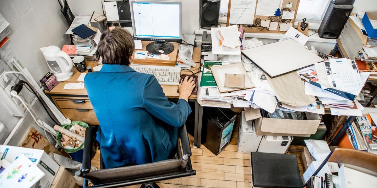 Uma mulher trabalha em seu escritório pessoal em uma mesa com pilhas de folhas e documentos que se espalham por todo o ambiente